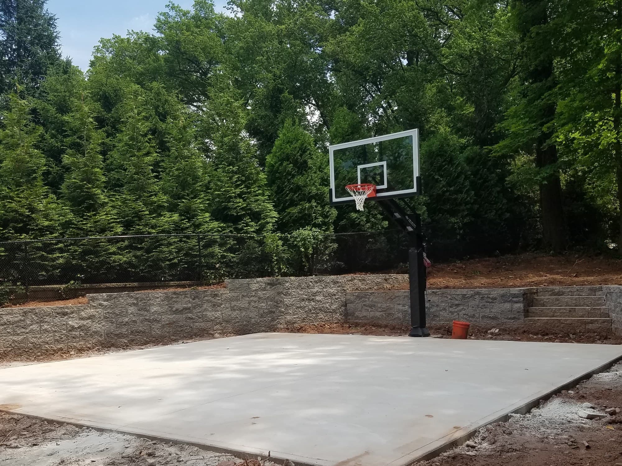 Concrete sport court with a basketball hoop in a side yard above a low retaining wall