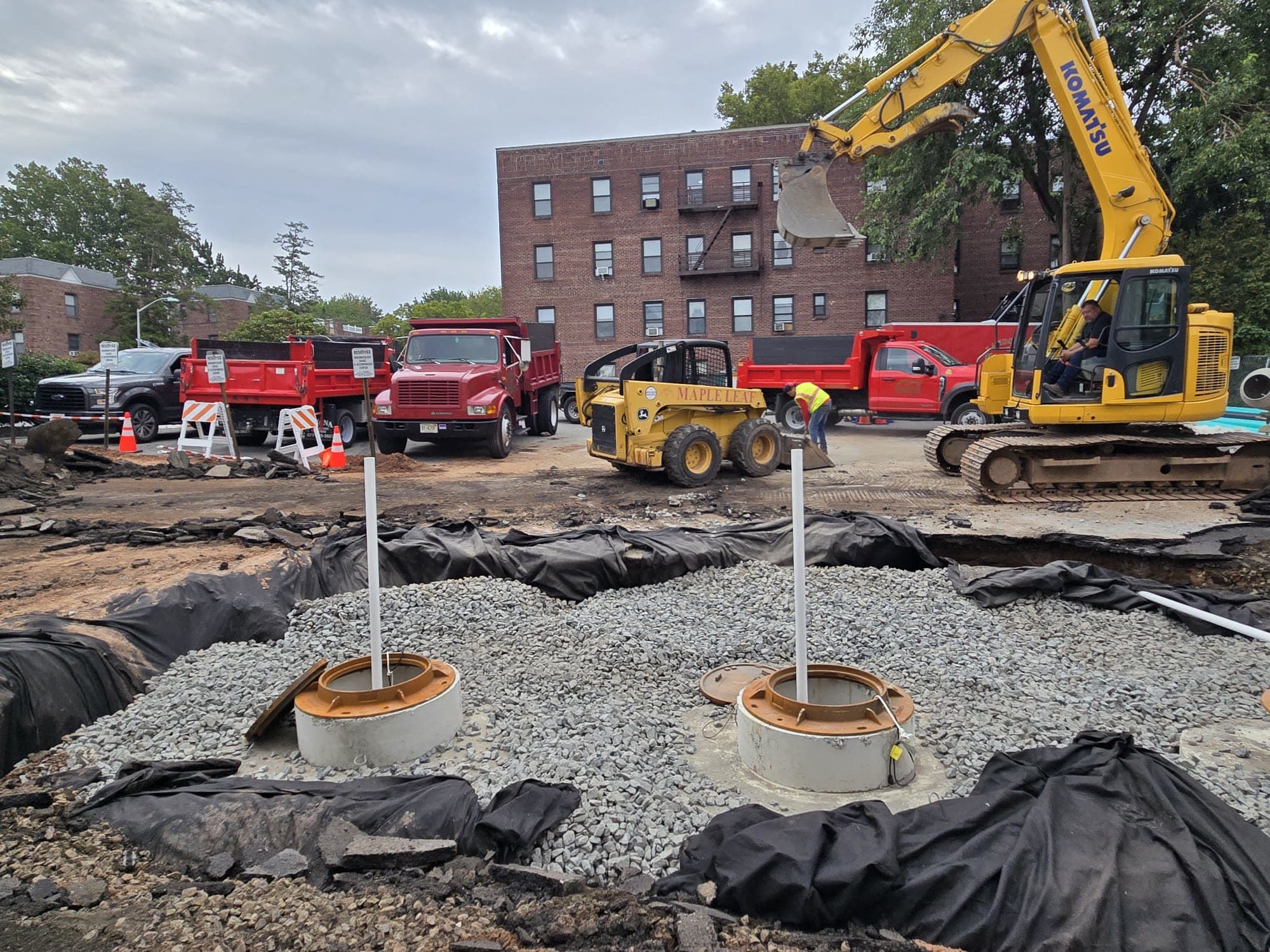 Two concrete seepage pits set in a fabric-lined stone bed outside an apartment building, excavator and crew trucks working behind