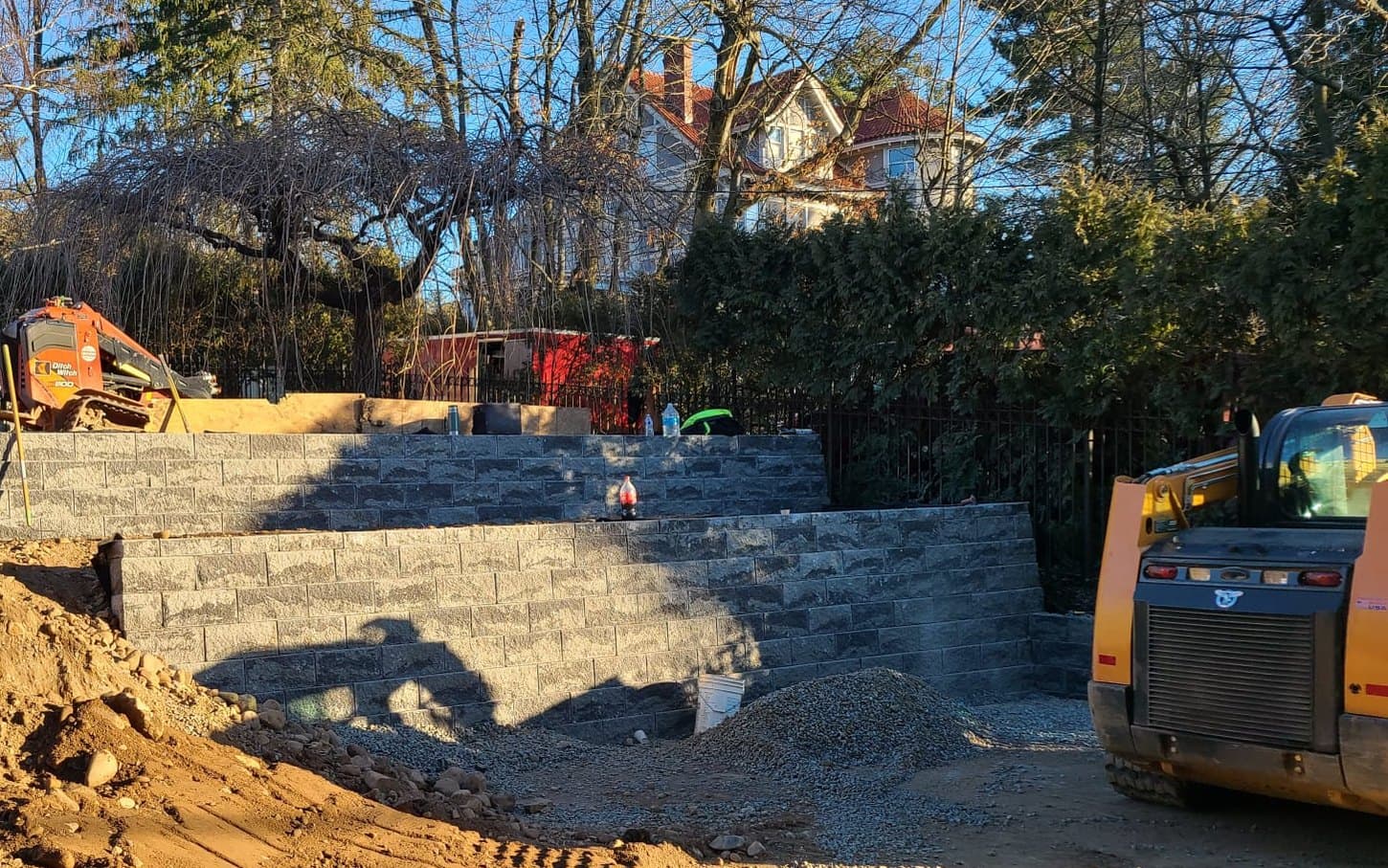 Two tiers of block retaining wall terracing a backyard slope, skid steer working behind