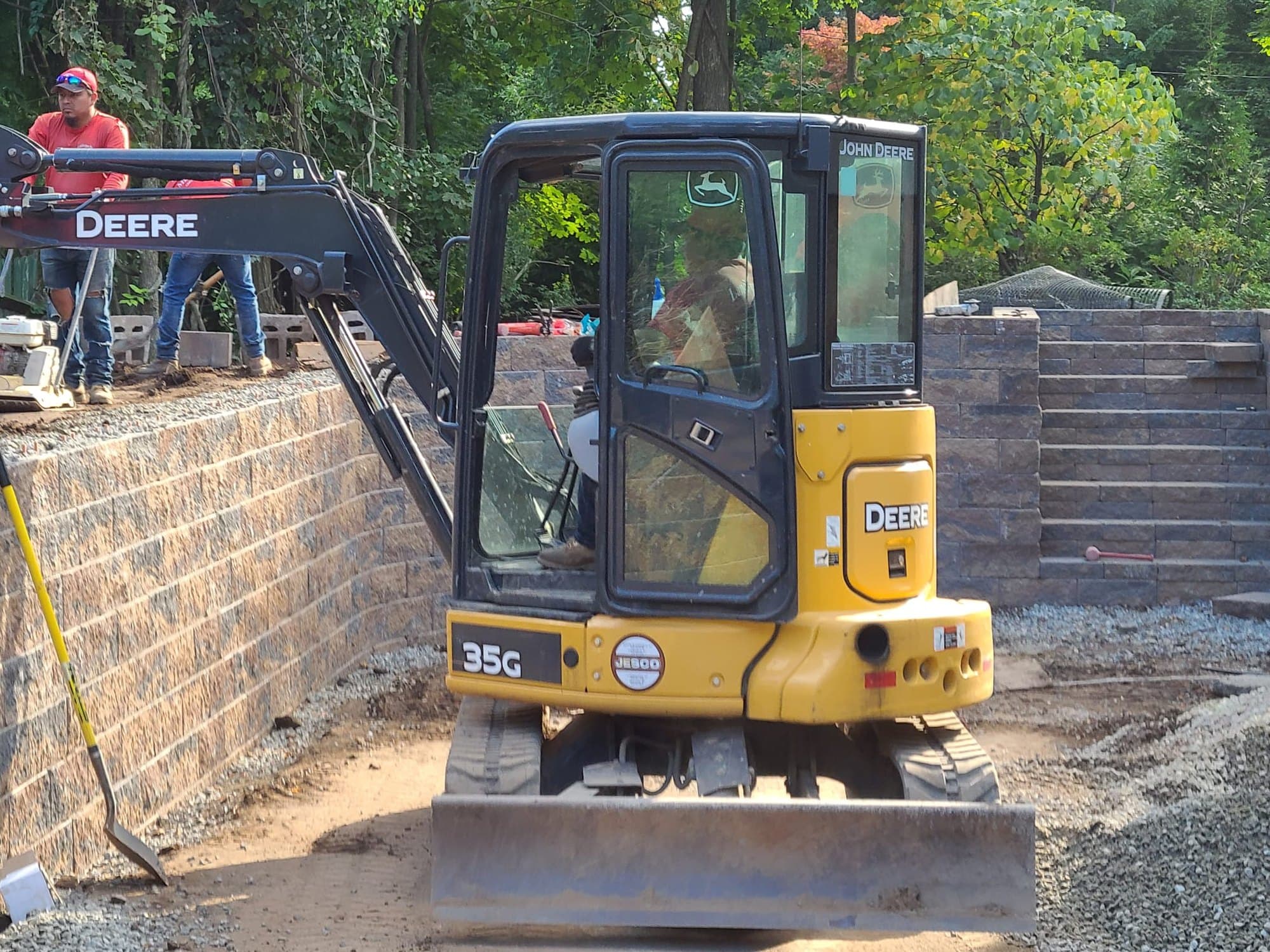 Excavator working on top of a new block retaining wall, with a second wall and steps behind